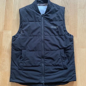 Svart dunväst från Les Deux - Stilren svart dunväst från Les Deux med diskret logga på bröstet. Västen har quiltad design, dragkedja framtill och två stora fickor. Perfekt för lager-på-lager och enkel att matcha med olika outfits.