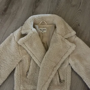 Beige teddyjacka från NA-KD - Supermjuk och fluffig teddyjacka från NA-KD i beige färg. Jackan har bred krage, dragkedjor framtill. Perfekt för dig som vill ha en trendig och cozy look under kyliga dagar.