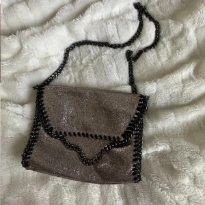 Glittrig axelväska med kedjedetaljer - Snygg glittrig axelväska i mörk silver med svarta kedjor längs kanterna och som axelrem. Väskan har en mjuk, fyrkantig form och lockstängning. Perfekt för dig som vill sticka ut med en trendig accessoar.