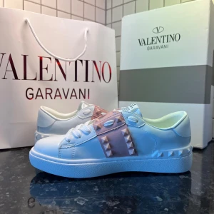 Valentino Rockstud sneakers vit/rosa - Snygga Valentino Garavani Rockstud sneakers i vitt skinn med ljusrosa band och ikoniska nitar. Klassisk rund tå, platt sula och vita snören. Märkeslogga på plösen och detaljerade nitar längs sidorna och sulan. Perfekta för dig som vill sticka ut med exklusiv streetstyle.