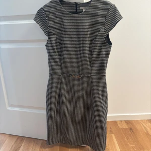 Rutig kortklänning från H&M - Snygg kortklänning från H&M i svartvitt rutigt mönster med guldfärgad detalj i midjan. Klänningen har rund hals, korta ärmar och figurnära passform. Perfekt för dig som gillar klassisk stil med en twist.