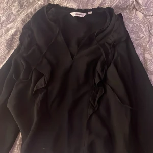 Svart volang blus  från Kappahl - Supersnygg svart blus  från Kappahl i storlek S. Blusen  har långa ärmar med resår vid ärmslut, v-ringning och fina volangdetaljer framtill. Tillverkad i ett lätt och mjukt polyestertyg som ger ett elegant fall.