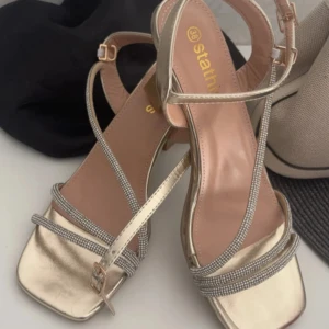Guldiga sandaler med strass och klack - Säljer ett par snygga sandaler i guld med smala remmar dekorerade med strass. Skorna har fyrkantig tå, öppen häl och en stabil klack. Perfekta för dig som vill sticka ut med glittriga detaljer och elegant design.