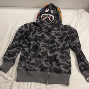 Bape hoodie - Säljer en ikonisk BAPE Shark hoodie i grått camomönster med hel dragkedja och broderade detaljer på huvan. Färgglada bokstäver och hajmotiv gör den riktigt unik. Perfekt streetwear-plagg med ribbade muddar och fickor framtill. Jag kan inte garantera äktheten på denna vara