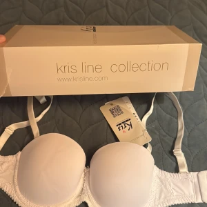 Vit balconette BH Kris Line 70B - Säljer en vit balconette BH från Kris Line, modell AfrodytA. Den har släta kupor, broderade spetsdetaljer på sidorna och justerbara axelband. Medföljer även genomskinliga axelband för extra flexibilitet. Perfekt under toppar med bar axel.