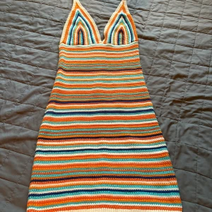 Randig virkad klänning från Ginatricot  - Färgglad virkad kortklänning från Ginatricot med smala axelband och djup v-ringning. Klänningen har ränder i orange, blått, turkost, vitt och beige. Perfekt för sommarens alla tillfällen och ger en riktigt retrovibe.