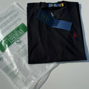 Svart t-shirt från Polo Ralph Lauren - Oanvänd svart t-shirt från Polo Ralph Lauren med klassisk röd broderad logga på bröstet. Modellen är kortärmad och har en rund halsringning. Tillverkad i mjuk bomull för en skön känsla. Perfekt för en clean och stilren look.