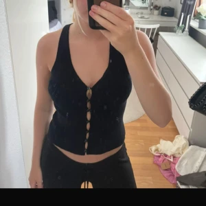 Svart ribbad linnetopp med knappar - Säljer en svart ribbad linnetopp med djup v-ringning och dekorativa knappar framtill. Toppen är ärmlös och har en croppad passform som ger en trendig look. Perfekt att matcha med jeans eller kjol för en cool stil. Lite i storleken. Är egentligen en xs/s i topp.