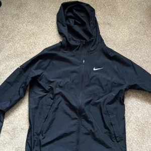Svart Nike Miller vindjacka med huva - Säljer en svart vindjacka från Nike med huva och dragkedja framtill. Jackan har två sidofickor med dragkedja och en vit Nike-logga på bröstet. Tillverkad i lätt polyester, perfekt för blåsiga dagar. Snygg och sportig design som funkar året runt.