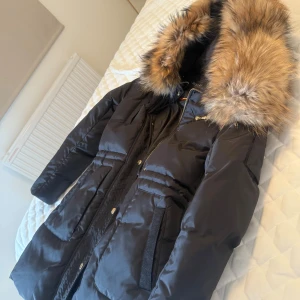 Svart pufferjacka med fuskpäls från Hollies - Svart pufferjacka från Hollies med stor huva kantad av fluffig fuskpäls i bruna toner. Jackan har dragkedja, knappar och flera praktiska fickor. Perfekt för kalla dagar med sin rejäla vaddering och snygga detaljer i guld.