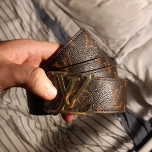 Louis Vuitton brunt skärp med LV-spänne - Säljer ett brunt Louis Vuitton-bälte i skinn med klassiskt monogrammönster och ett stort LV-spänne i guld. Bältet har exklusiv känsla och detaljerad finish, perfekt för dig som vill sticka ut med en ikonisk accessoar.