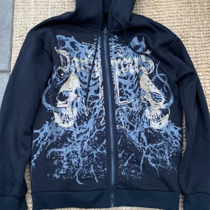 Svart hoodie med döskalletryck från Romwe - Svart hoodie från Romwe i storlek M med dragkedja framtill och huva. Framsidan har ett stort tryck med döskallar. Tröjan har långa ärmar och ribbade muddar. Perfekt för dig som gillar edgy streetstyle.