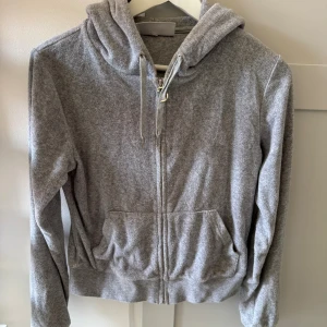 Grå hoodie från Juicy Couture - Mysig grå hoodie från Juicy Couture i frotté med dragkedja och huva. Tröjan har snörning i halsen, två fickor fram. Storlek S