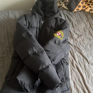 Svart pufferjacka Zavetti Canada XS - Svart pufferjacka från Zavetti Canada med huva och patch på ärmen. Jackan har dragkedja, tryckknappar och flera fickor. Insidan är fodrad med mjukt material och logotypdetaljer. Perfekt för kalla dagar när du vill hålla stilen.