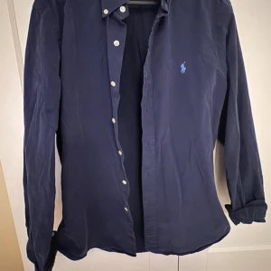 Mörkblå skjorta från Ralph Lauren - Snygg mörkblå skjorta från Ralph Lauren i slim fit-modell. Skjortan har klassisk krage, knäppning framtill med vita knappar och broderad logga på bröstet. Tillverkad i bomull och har långa ärmar med manschettknappar.