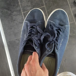 Mörkblå sneakers från Tommy Hilfiger - Snygga mörkblå sneakers från Tommy Hilfiger med klassisk vit sula och diskret logga på sidan. Skorna har rund tå, snörning och är tillverkade i textilmaterial. Perfekta för en stilren och avslappnad look.