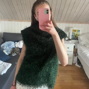 Mörkgrön fluffig stickad väst - Säljer en riktigt cool mörkgrön stickad väst med fluffig och lite glittrig känsla. Västen har bred krage och är ärmlös, perfekt för att ge din outfit en unik touch. Passar dig som gillar att sticka ut och vill ha något annorlunda i garderoben.