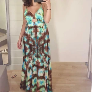 Maxi klänning - Säljer en maxiklänning med batikmönster i blått, brunt och grönt. Klänningen har smala axelband, djup v-ringning och en snygg cut-out detalj framtill. Perfekt för sommardagar och festivaler. Materialet känns lätt och flowy.