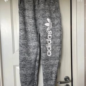 Gråa mjukisbyxor från Adidas - Gråmelerade mjukisbyxor från Adidas med stort vitt tryck och logga längs ena benet. Byxorna har resår i midjan och vid bensluten, samt en avslappnad passform som är perfekt för chill dagar.