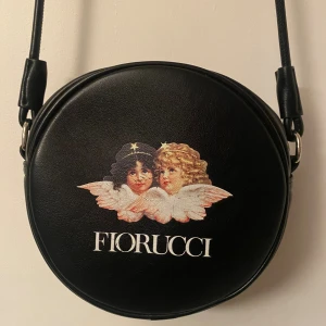 Svart rund axelväska från Fiorucci - Cool svart rund axelväska från Fiorucci med ikoniskt ängeltryck framtill och vit logga. Väskan har lång axelrem och är gjord i skinnimitation. Perfekt för dig som vill sticka ut med en unik accessoar.