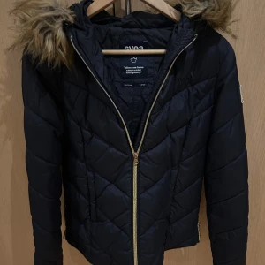 Svart pufferjacka med fuskpäls från Svea - Säljer en svart pufferjacka från Svea i storlek XS. Jackan har huva med fuskpäls, guldig dragkedja och quiltad design. Perfekt för kalla dagar och riktigt snygg till jeans eller leggings. Broderad Svea-logga på bröstet och patch på ärmen.