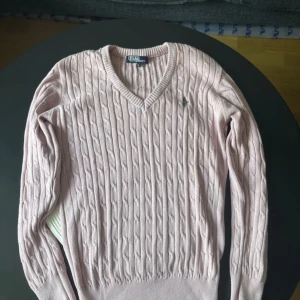 Ljusrosa kabelstickad tröja Polo Ralph Lauren - Supersnygg ljusrosa kabelstickad tröja från Polo Ralph Lauren med v-ringning och broderad logga på bröstet. Tröjan har lång ärm och är gjord i mjuk bomull, perfekt för lager-på-lager under höst och vår.