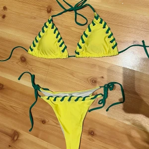 Gul bikini med gröna snörningar - Snygg gul bikini med gröna snörningsdetaljer längs kanterna. Triangeltopp med knyt i nacken och ryggen samt matchande bikiniunderdel med knyt i sidorna. Perfekt för stranden eller poolen i sommar! Använts 1 gång.