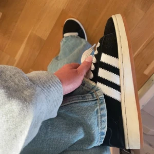 Adidas Campus svarta sneakers mocka - Svarta Adidas Campus sneakers i mocka med klassiska vita ränder på sidorna och vit sula med gummiyta. Skorna har breda vita snören och rund tå. Perfekta för en chill streetstyle-look och funkar året runt