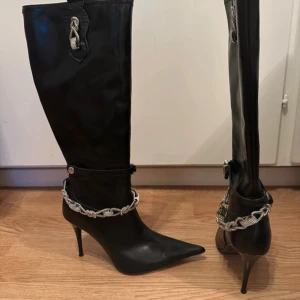 Svarta höga stövlar med klack från Bianco - Snygga svarta stövlar med spetsig tå och hög klack. Dekorerade med en glittrig silverfärgad kedja runt ankeln och ett matchande silverhänge upptill. Perfekta för dig som vill sticka ut med detaljer och elegant vibe. Klacken är 10cm