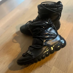 Svarta moon boots med snörning - Säljer ett par svarta moon boots med glansig finish och vadderad ovandel. Skorna har rund tå, kraftig platt sula och snörning med metallspännen som går runt hela skon. Perfekta för vintern och riktigt sköna att ha på sig när det är kallt.
