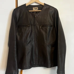 Brun läderjacka i storlek 40 med märket Joy - Stilren brun läderjacka med rund halsringning och raka linjer. Jackan har två bröstfickor med dragkedja och hel dragkedja framtill. Tillverkad i mjukt skinn med slät insida, perfekt för dig som gillar en clean look.