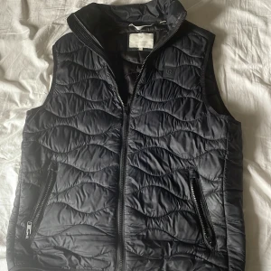 Svart quiltad dunväst Jack & Jones - Svart quiltad dunväst från Jack & Jones med vågigt mönster i tyget. Västen har hög krage, två fickor med dragkedja och hel dragkedja framtill. Perfekt för lager-på-lager och snygg till både hoodie och t-shirt.