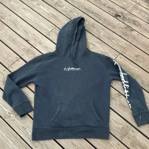 Svart Hollister hoodie med tryck - Svart  hoodie från Hollister med vit logga på bröstet och stort tryck längs ena ärmen. Tröjan har huva med snörning och en klassisk magficka. Perfekt för chill dagar och enkel att matcha med jeans eller joggers.