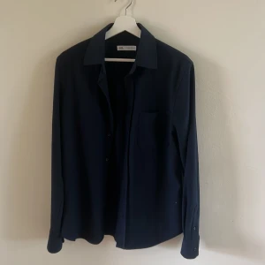 Mörkblå skjorta från Zara - Snygg mörkblå skjorta från Zara med klassisk krage och bröstficka. Skjortan har lång ärm och knappar framtill. Perfekt för dig som gillar stilrena och enkla plagg. Materialet känns mjukt och lätt mot huden. Kan även användas som overshirt.