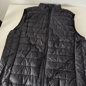 Svart quiltad dunväst med dragkedja - Säljer en svart dunväst med quiltad design och hög krage. Västen har en hel dragkedja framtill och är lätt vadderad, perfekt för lager-på-lager. Enkel och stilren modell som funkar till många outfits. Helt ny.