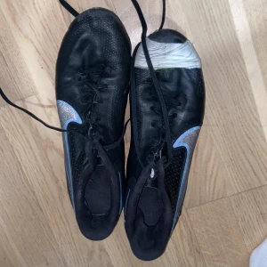 Nike Tiempo svarta fotbollsskor - Svarta Nike Tiempo fotbollsskor med silvriga detaljer och snörning. Skorna har klassisk rund tå och platt sula med fasta dobbar för bra grepp på planen. Tillverkade i skinn och syntetmaterial, med Nike-logga på sidan och Tiempo-tryck under sulan.