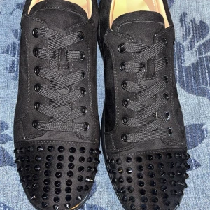 Christian Louboutin svarta sneakers med nitar - Svarta sneakers från Christian Louboutin i mocka med coola svarta nitar på tån och klassisk röd sula. Skorna har snörning och en rund tå. Perfekt för dig som vill sticka ut med en lyxig och edgy stil.