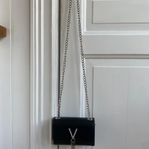 Svart axelväska från Mario Valentino - Snygg svart axelväska från Mario Valentino i skinn med silverfärgad kedja och blingad V-detalj framtill. Väskan har en elegant tofs i metallkedjor och stilren, kompakt form. Perfekt för dig som vill ha en trendig och lyxig accessoar.