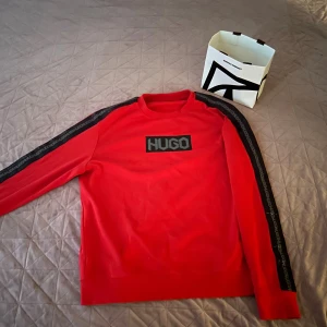 Röd HUGO sweatshirt med logga - Röd sweatshirt från HUGO med svart logga framtill och svarta band med HUGO-tryck längs ärmarna. Klassisk rund hals och långa ärmar. Skön och stilren tröja i bomull, perfekt för en trendig streetstyle-look.
