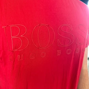 Röd Hugo Boss t-shirt med tryck - Snygg röd t-shirt från Hugo Boss med stort BOSS-tryck på bröstet och diskret logga i nacken. Klassisk rund hals och korta ärmar. Mjuk bomullskvalitet som känns skön mot huden. Perfekt för dig som gillar stilrena logotoplagg.