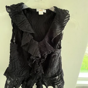 Svart volangblus från Gina Tricot - Säljer en svart ärmlös blus från Gina Tricot med volangdetaljer längs v-ringningen och nederkanten. Tunt, lätt transparent tyg med vågig struktur och knytband framtill. Perfekt för dig som gillar en edgy och romantisk stil.