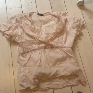 Ljusrosa blus med volang från Wet Seal - Söt ljusrosa blus från Wet Seal med korta puffärmar och v-ringning med volangdetalj. Blusen har resår i midjan för en feminin siluett och är tillverkad i ett tunt, lätt material som passar perfekt till vår och sommar.