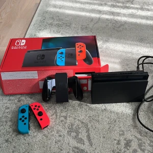 Nintendo Switch - Nintendo Switch med neonröda och neonblå Joy-Con, originalkartong, dockningsstation och tillbehör. Konsolen och kontrollerna ser ut att vara i mycket gott skick utan synliga repor eller skador. Perfekt för både bärbart och stationärt spelande.Om du går in på mitt konto finns det en massa spel du kan välja till Nintendo. Det finns en extra Nintendo Switch som jag köpte vilket är bra om du vill spela med fler vänner, och det finns också en riktigt bra Nintendo-väska där du kan lägga Switchen.