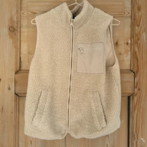 Beige fleeceväst med dragkedja - Mysig beige fleeceväst med hög krage och hel dragkedja framtill. Västen har två sidofickor och en bröstficka med dragkedja i matchande färg. Perfekt för lager-på-lager och chill dagar. Snygg och enkel att styla till det mesta.