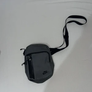 Grå axelväska från Nike - Snygg grå axelväska från Nike med justerbart svart band och flera fack med dragkedja. Väskan har en stilren design med Nike-logga framtill och är tillverkad i slitstark polyester. Perfekt storlek för att bära med sig det viktigaste.