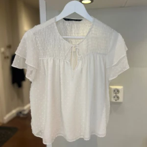 Vit prickig blus från Zara Basic - Verkligen sååå söt och fin! Använd Max 1 gång. Finare material. Storlek L men sitter som en M. Supersöt vit blus från Zara Basic med små prickar i tyget, volangdetaljer och knytning vid halsen. Blusen har korta, luftiga ärmar och smock upptill för en avslappnad och flowy känsla. Perfekt till jeans eller kjol för en fräsch look. 🥰🥰