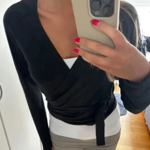 Svart omlottopp med knytband - Svart omlottopp med långa ärmar och knyt i sidan. Toppens omlottmodell ger en snygg v-ringning och markerad midja. Mjuk och stretchig känsla, perfekt att styla med jeans eller kjol. Köpt här på plick men har inte kommit till någon användning 💗💗💗