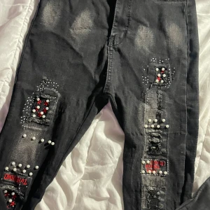 Svarta jeans med pärlor och broderi - Unika svarta jeans med slitna detaljer, pärlor i vitt och rött samt broderade lappar med text. Jeansen har en skinny passform och coola patchar framtill som ger en edgy vibe. Perfekta för dig som vill sticka ut med din stil.