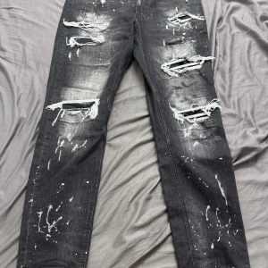 Svarta distressed jeans Supply & Demand - Svarta jeans från Supply & Demand med kraftiga slitningar och trasiga detaljer framtill. Byxorna har vit färgstänkseffekt och raka ben. Snyggt streetwear-plagg med coola slitningar och tvättad look. Passar dig som gillar en edgy stil.