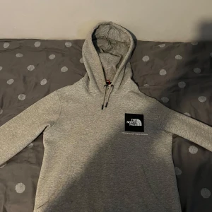 Grå hoodie från The North Face - Snygg grå hoodie från The North Face med stor huva och snörning. Tryck med logga och texten 'Never Stop Exploring' på bröstet. Mjuk och bekväm, perfekt för chill eller äventyr. Klassisk passform och långärmad modell.